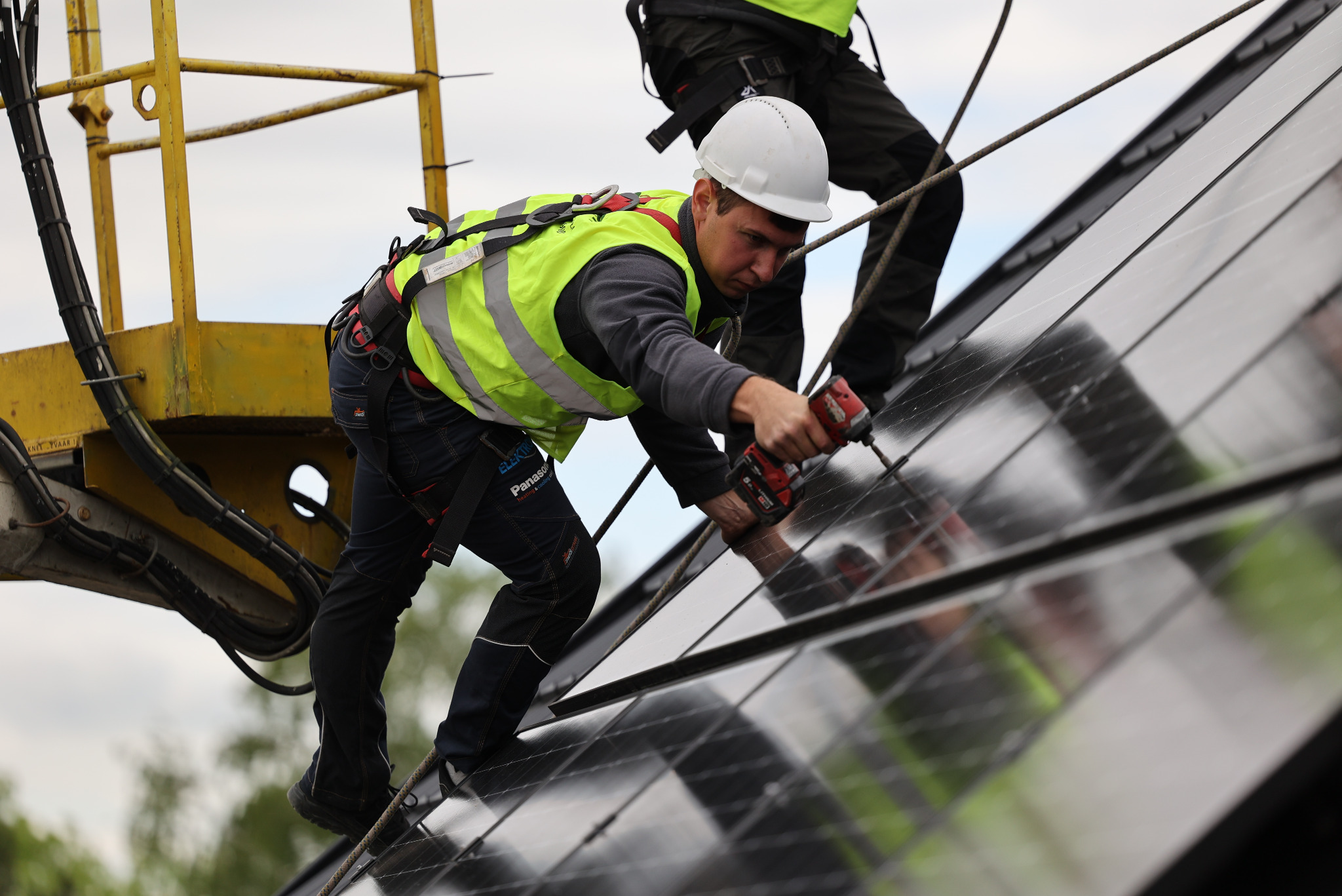 Instalator w kasku i kamizelce, z wiertarką, montuje panele fotowoltaiczne na dachu z platformy. Widoczne zabezpieczenia linowe i narzędzia.