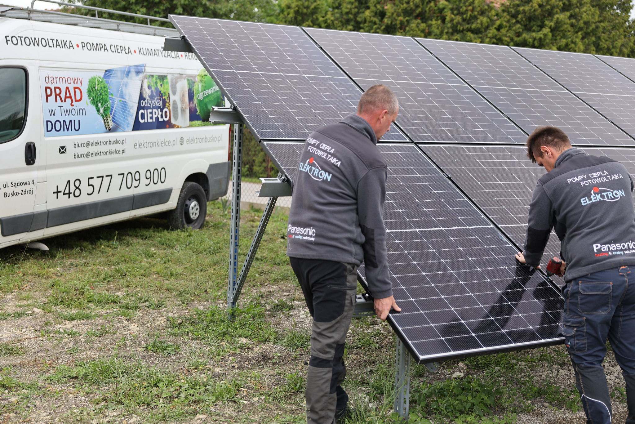 Montaż paneli fotowoltaicznych przez dwóch instalatorów w szarych bluzach z logo firmy, na tle busa z reklamą firmy i numerem telefonu.