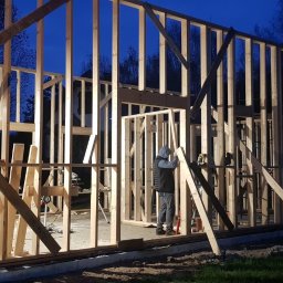 Timber House Tomasz Wojciechowski - Konstrukcja szkieletowa domu w trakcie budowy nocą, widoczny pracownik wewnątrz. Elementy drewniane tworzą ramy ścian i otworów okiennych.