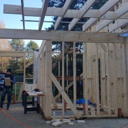 Timber House Tomasz Wojciechowski - Konstrukcja drewniana domu w trakcie budowy. Widoczny szkielet ścian, stropu i pracownik z deską na tle lasu i błękitnego nieba. Na pierwszym planie piła stołowa.