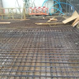 Timber House Tomasz Wojciechowski - Zbrojenie fundamentów domu jednorodzinnego, siatka z prętów stalowych ułożona na mokrej powierzchni, przygotowana do zalania betonem. Widoczne elementy szalunku.