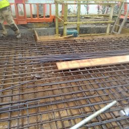 Timber House Tomasz Wojciechowski - Zbrojenie fundamentu domu z widoczną siatką stalową, deskowaniem i robotnikiem w odblaskowej kamizelce. Detal z placu budowy w trakcie prac.