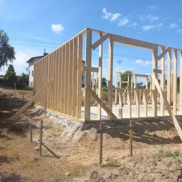 Timber House Tomasz Wojciechowski - Konstrukcja szkieletowa domu w trakcie budowy, widoczne drewniane ramy ścian i stropu na fundamencie, w tle błękitne niebo i częściowo zabudowa jednorodzinna.