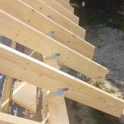 Timber House Tomasz Wojciechowski - Jasne drewniane krokwie dachu połączone metalowymi łącznikami, widoczne w zbliżeniu z góry, z wiórami na czarnej membranie dachowej i zielenią w tle.