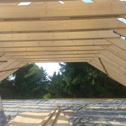Timber House Tomasz Wojciechowski - Konstrukcja dachu z jasnego drewna, widok od wewnątrz, z widocznymi belkami i deskami. W tle zieleń drzew przez otwór w dachu. Wióry na membranie dachowej.