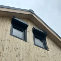 Fragment elewacji domu z jasnego drewna z dwoma oknami z czarnymi roletami zewnętrznymi. Widok na szary dach i pochmurne niebo.