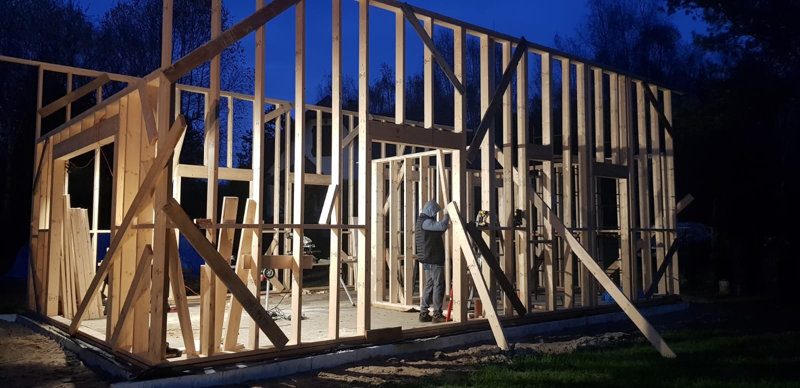 Konstrukcja szkieletowa domu w trakcie budowy nocą, widoczny pracownik wewnątrz. Elementy drewniane tworzą ramy ścian i otworów okiennych.