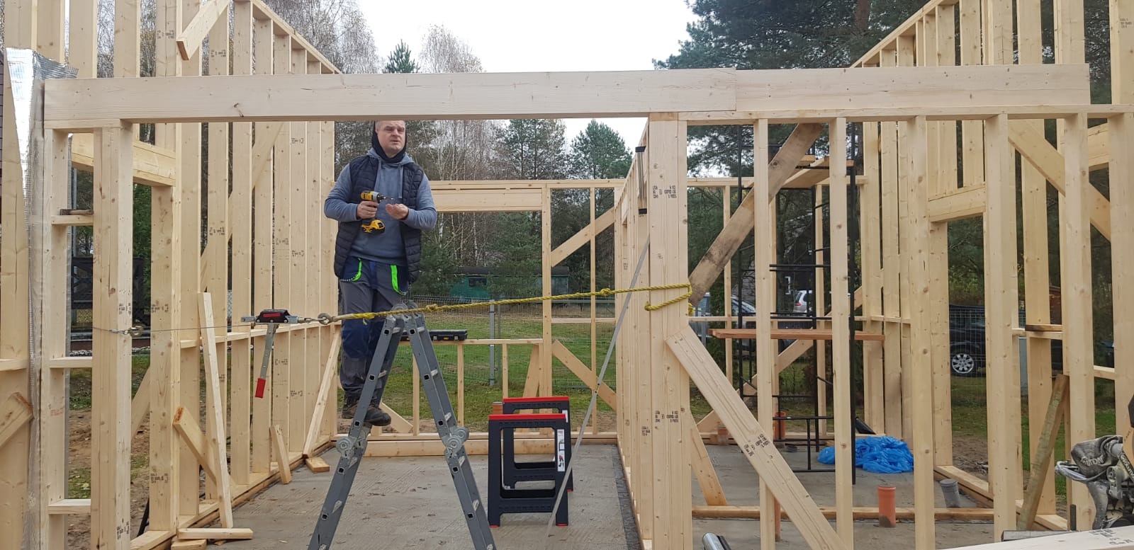 Budowa domu szkieletowego: mężczyzna na drabinie z wiertarką montuje drewnianą konstrukcję ścian. Widoczne narzędzia i elementy konstrukcyjne. Tło: drzewa.