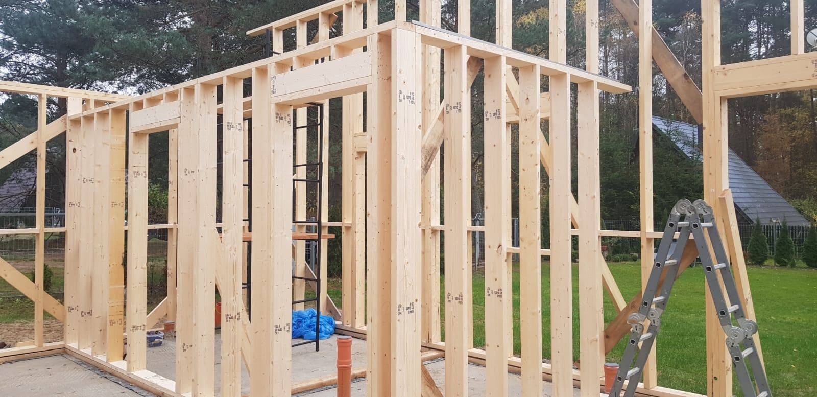 Konstrukcja szkieletowa domu w trakcie budowy, widoczne drewniane elementy ścian, słupy i belki. Na zewnątrz trawa i drzewa, w tle dom z ciemnym dachem.