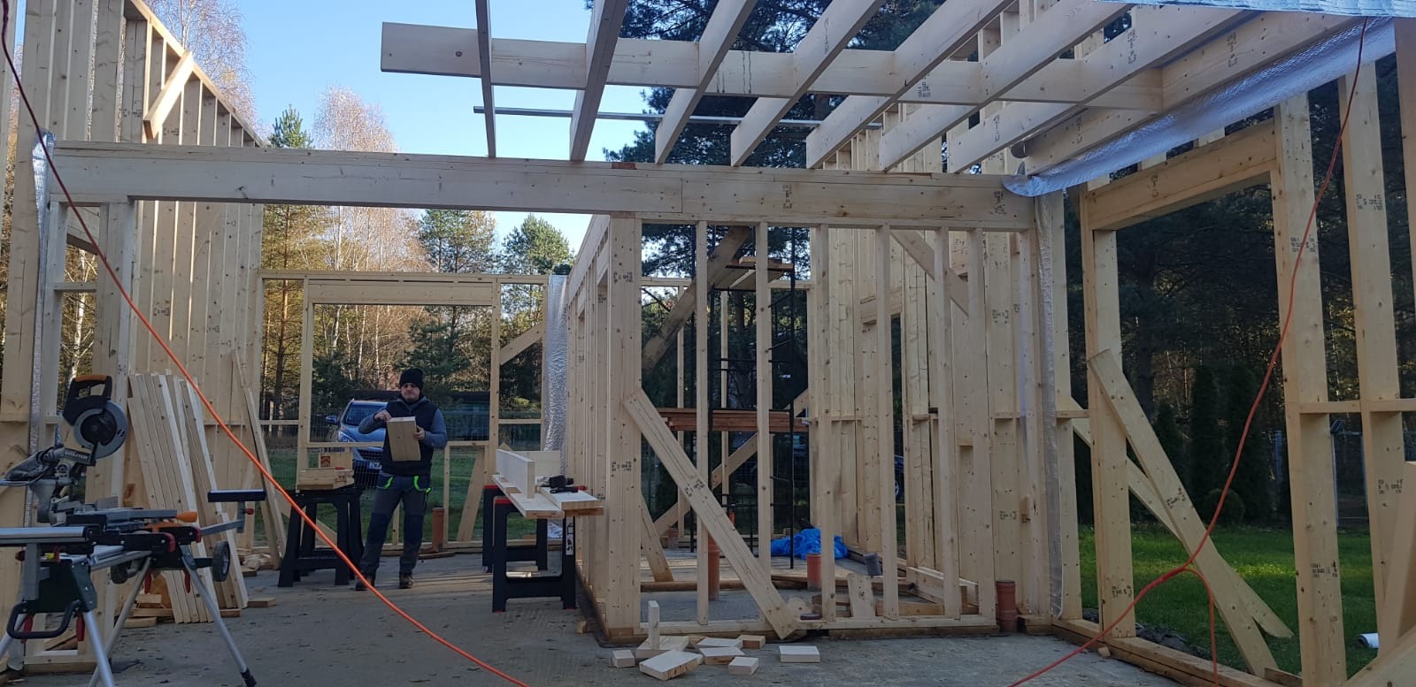 Konstrukcja drewniana domu w trakcie budowy. Widoczny szkielet ścian, stropu i pracownik z deską na tle lasu i błękitnego nieba. Na pierwszym planie piła stołowa.