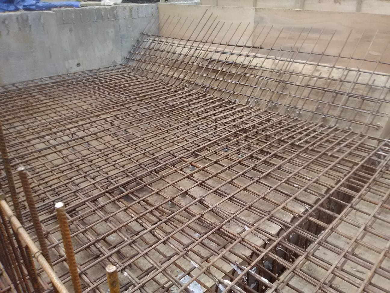 Zbrojenie fundamentów w formie siatki stalowej, widoczne pręty pionowe na krawędzi. Betonowe ściany szalunkowe w tle, gotowe do zalania betonem.