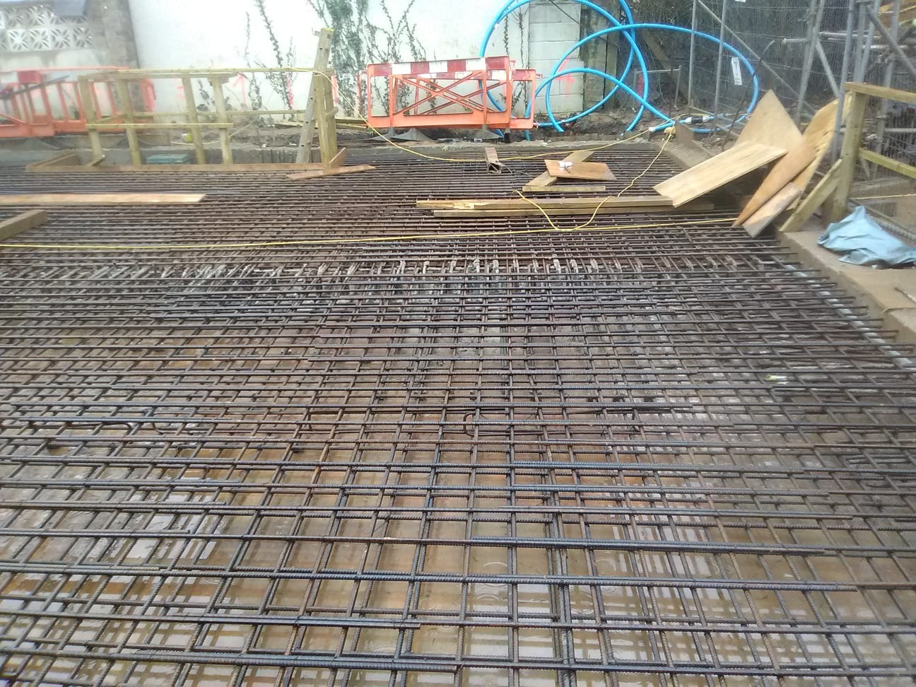 Zbrojenie fundamentów domu jednorodzinnego, siatka z prętów stalowych ułożona na mokrej powierzchni, przygotowana do zalania betonem. Widoczne elementy szalunku.