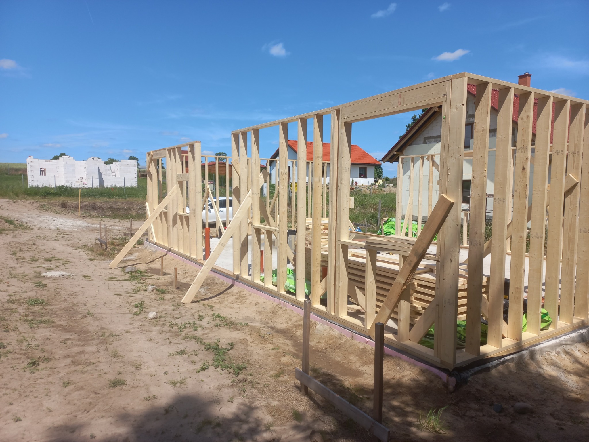 Wznoszenie konstrukcji szkieletowej domu jednorodzinnego. Widoczne drewniane ramy ścian na fundamencie, w tle budynek w trakcie budowy i błękitne niebo.