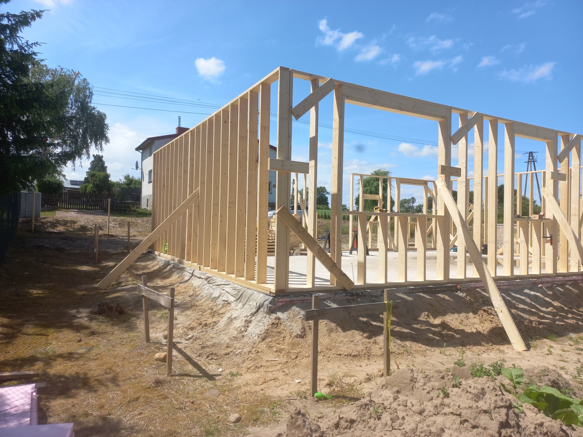 Konstrukcja szkieletowa domu w trakcie budowy, widoczne drewniane ramy ścian i stropu na fundamencie, w tle błękitne niebo i częściowo zabudowa jednorodzinna.