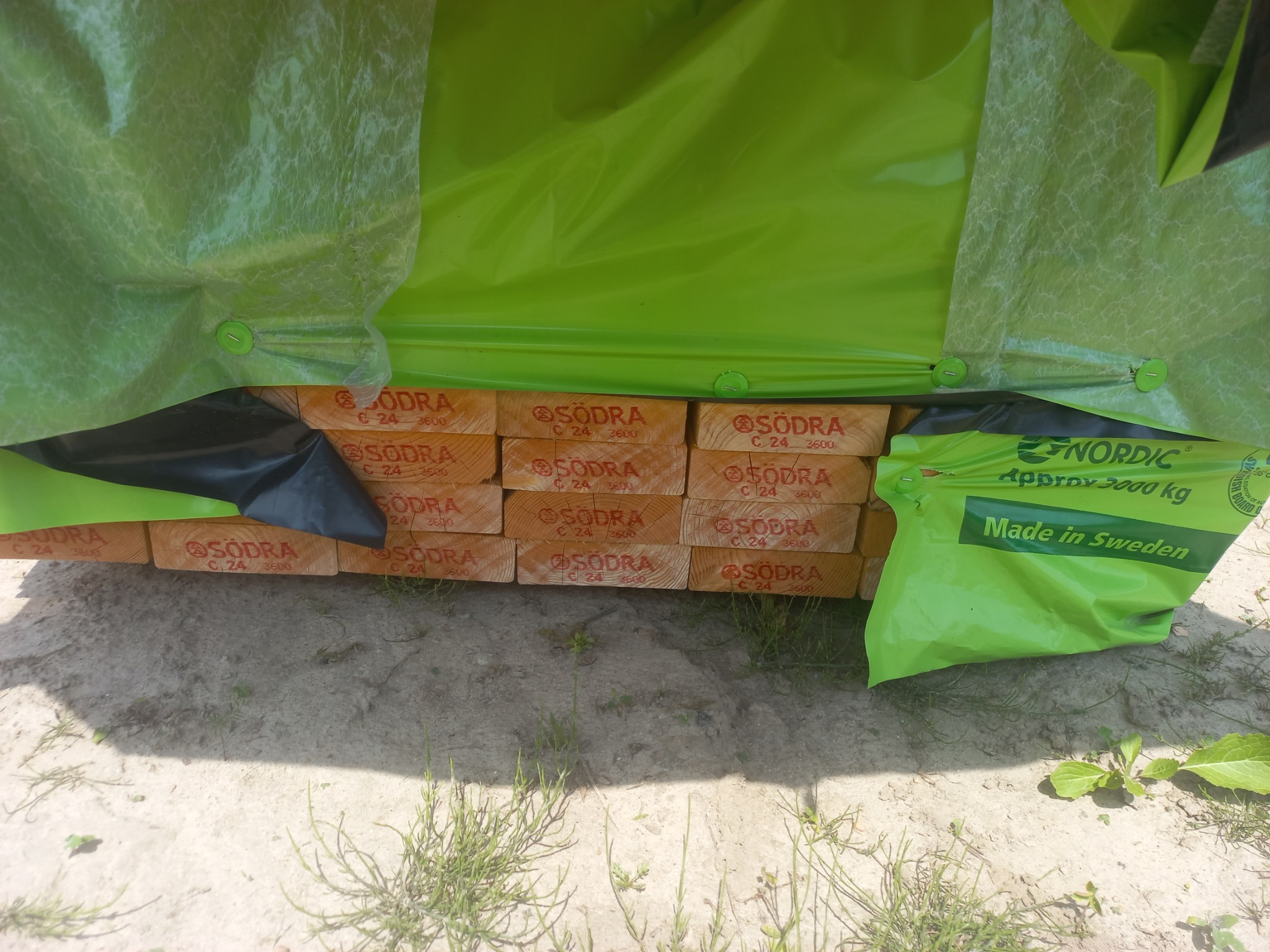 Stos belek drewnianych marki Södra C24 3600, częściowo przykrytych zieloną folią ochronną z napisem Nordic, leżących na piaszczystym podłożu z roślinnością.