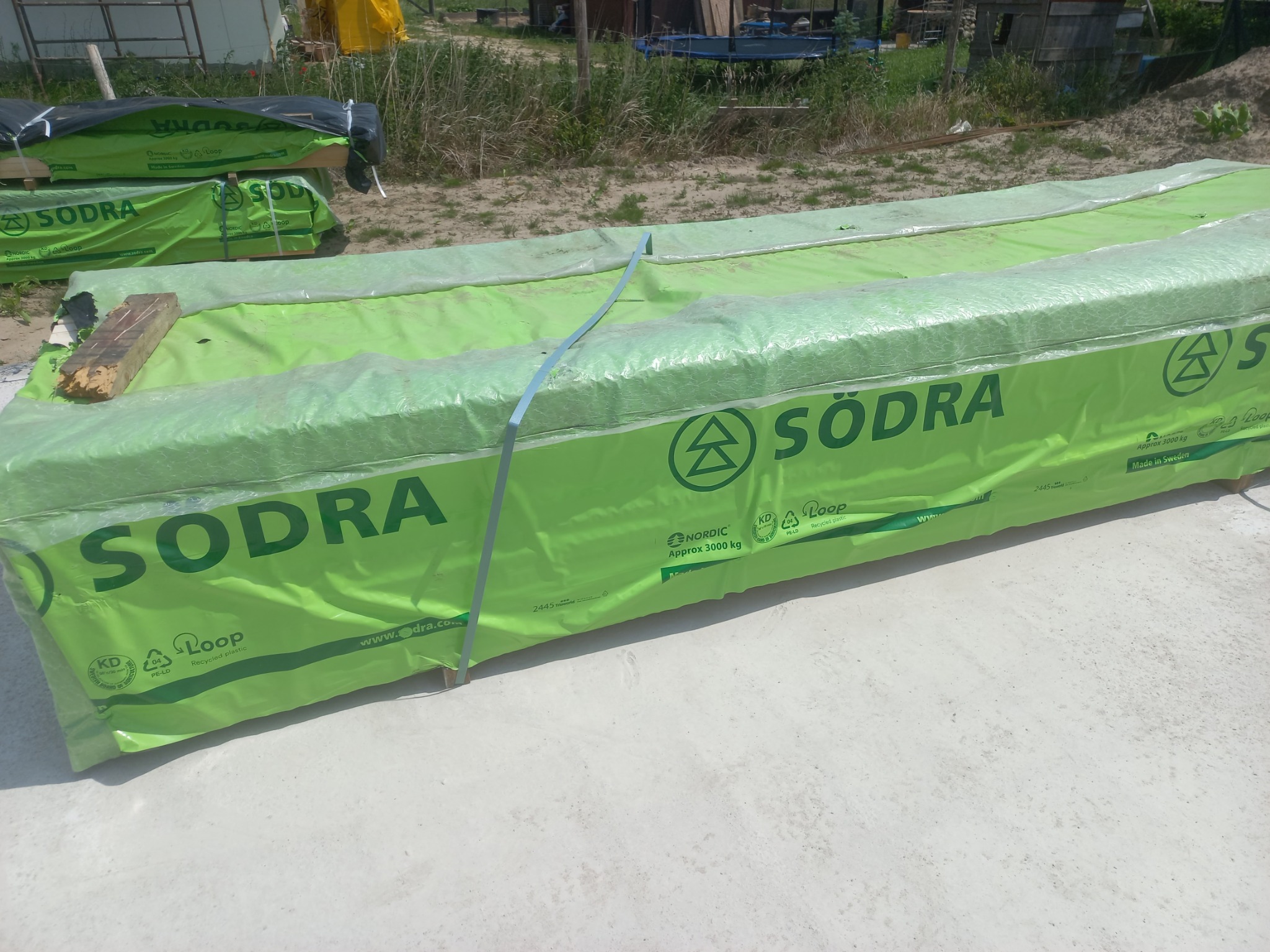 Paczka drewna konstrukcyjnego Södra w zielonej folii, zabezpieczona taśmą, leżąca na betonowym podłożu. W tle widoczne elementy konstrukcji budowlanej.