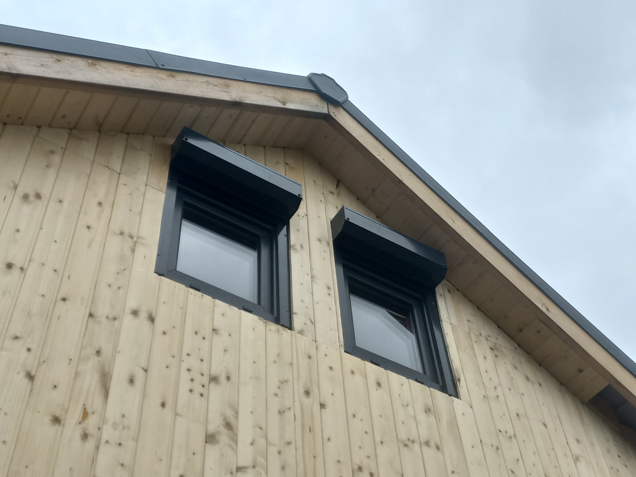 Fragment elewacji domu z jasnego drewna z dwoma oknami z czarnymi roletami zewnętrznymi. Widok na szary dach i pochmurne niebo.
