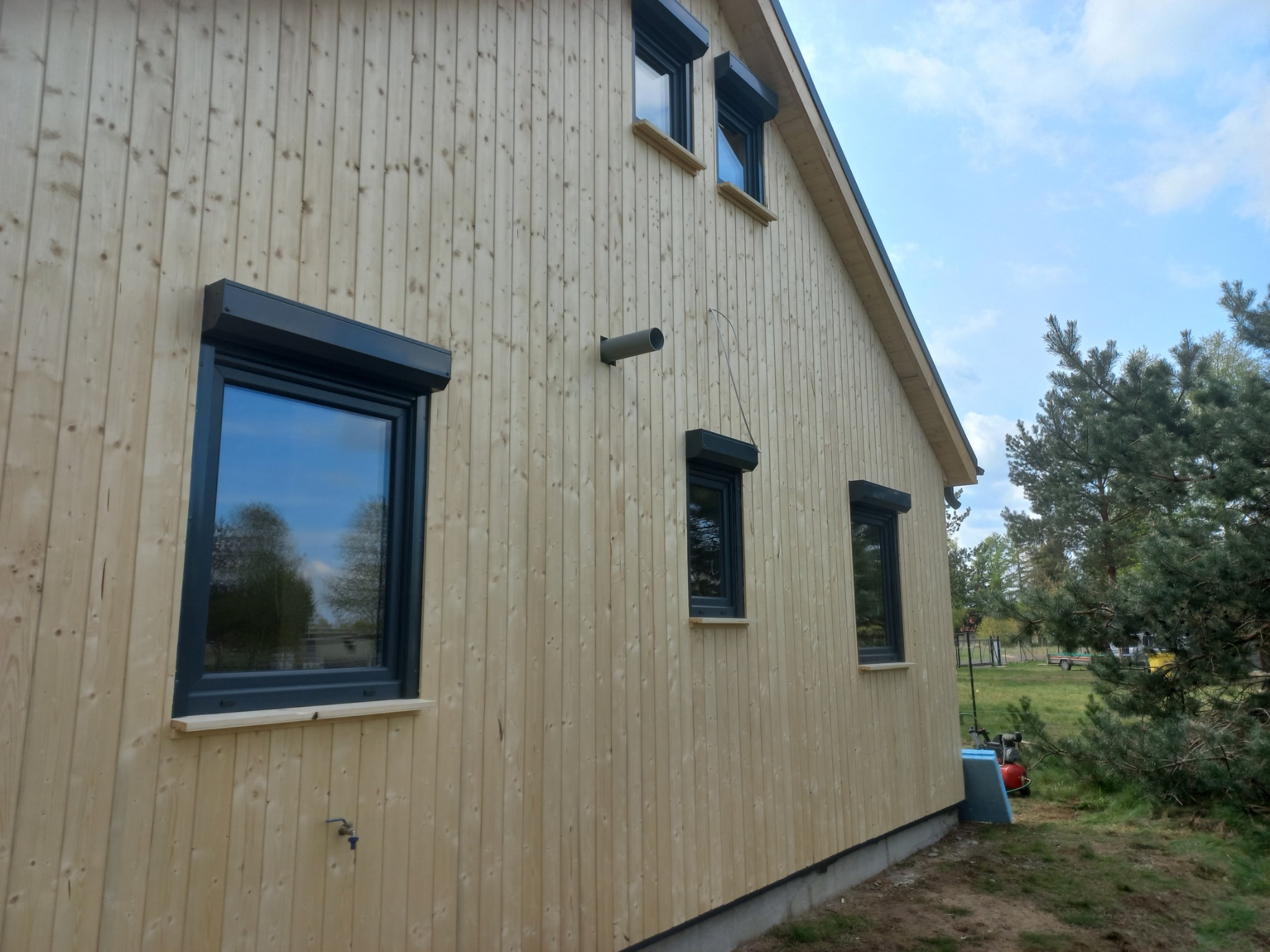 Elewacja domu szkieletowego z jasnego drewna, okna z roletami zewnętrznymi. Widoczny fragment ogrodu i błękitne niebo.