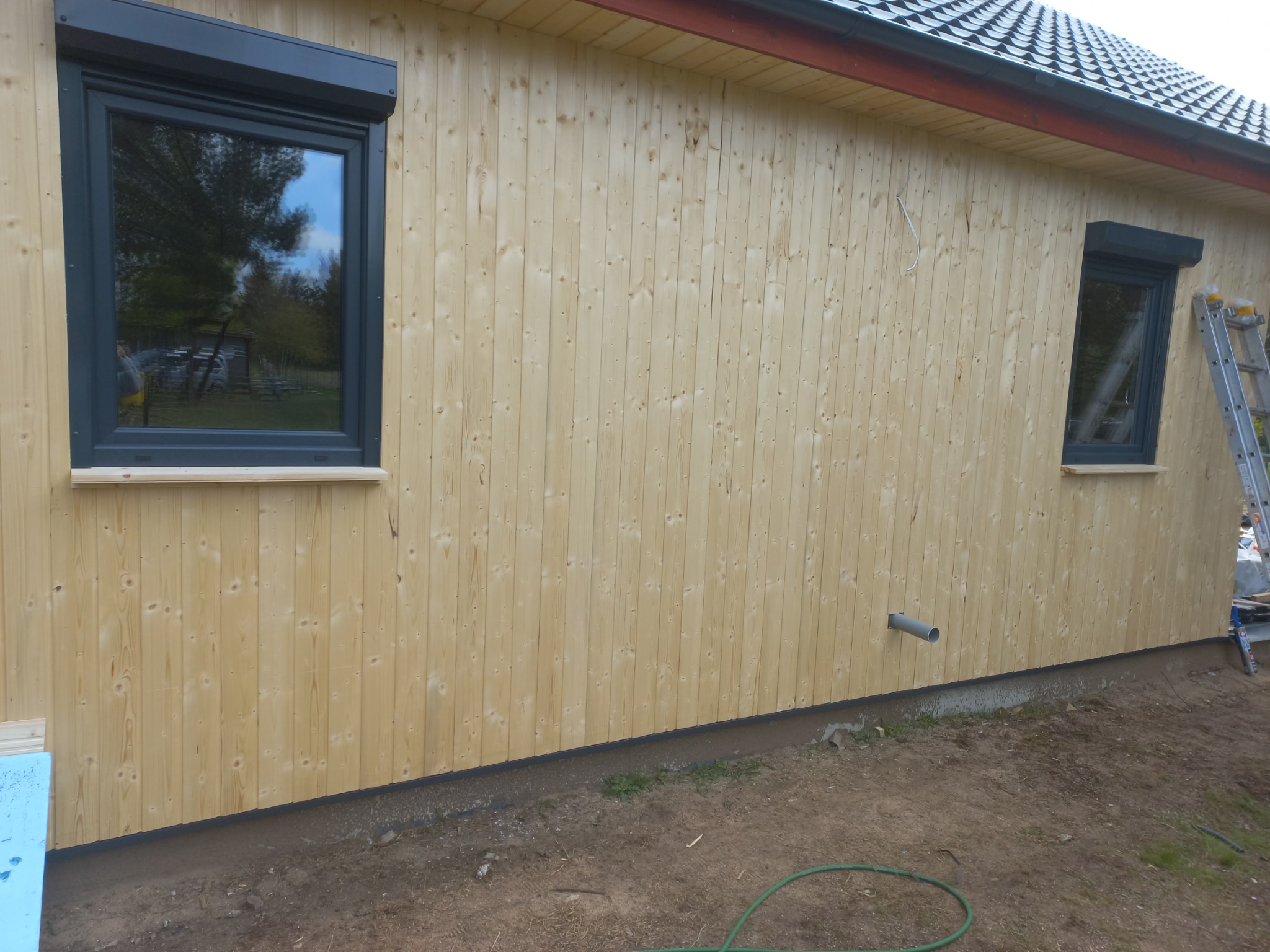 Ściana domu z jasnego drewna z dwoma oknami z szarymi ramami i roletami. Na ziemi widoczny wąż ogrodowy. Dom z zewnątrz.