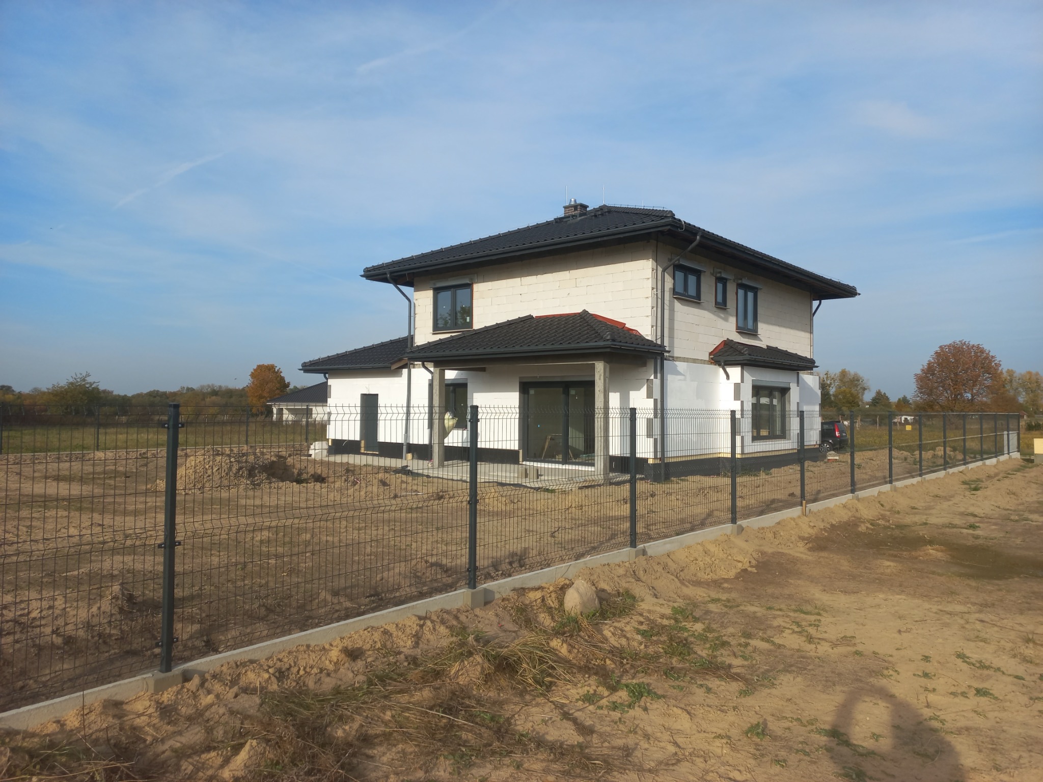 Dwukondygnacyjny dom w trakcie budowy, ogrodzony siatką. Widoczne pustaki, czarna dachówka i fragmenty izolacji. Tło: błękitne niebo i drzewa.