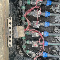 Ekspert ogrodu - Złożony system nawadniania w czarnej skrzynce z rurami, zaworami i okablowaniem, otoczony drobnymi kamieniami. Widoczne zielone złączki i niebieskie nakrętki na rurach.