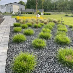 Nowoczesny ogród z ozdobnymi trawami i szarą korą kamienną, oświetlony kulistymi lampami. Widok na uporządkowaną przestrzeń z elementami architektury.