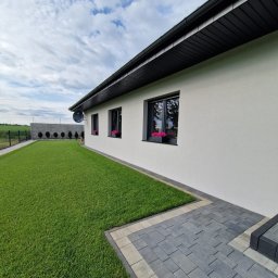 Ekspert ogrodu - Nowoczesny dom z równo przystrzyżonym trawnikiem, betonowym ogrodzeniem i szarą kostką brukową. W oknach widoczne różowe kwiaty w doniczkach.