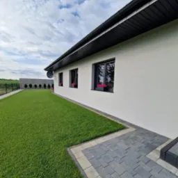 Nowoczesny dom z równo przystrzyżonym trawnikiem, betonowym ogrodzeniem i szarą kostką brukową. W oknach widoczne różowe kwiaty w doniczkach.