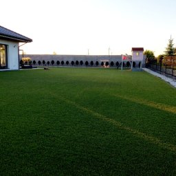 Ekspert ogrodu - Rozległy trawnik z rolki przed domem z szarym ogrodzeniem panelowym i ozdobnymi drzewkami. W tle widoczny domek dla dzieci i robot koszący.