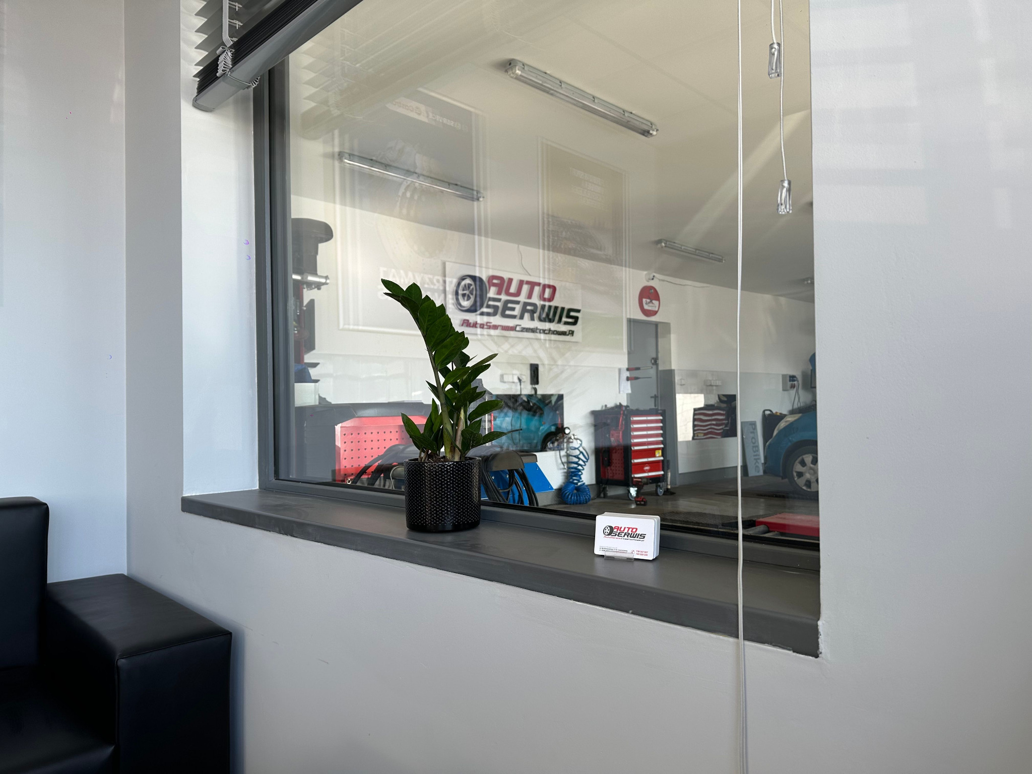 Poczekalnia warsztatu samochodowego z widokiem na stanowiska naprawcze przez okno. Na parapecie roślina w doniczce i wizytówka.