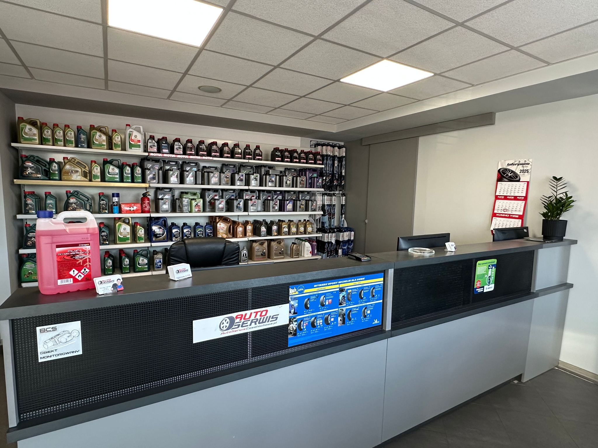 Wnętrze auto serwisu: lada recepcyjna z logo, półki z olejami silnikowymi, kalendarz Pirelli 2025. Widok na stanowisko obsługi klienta.