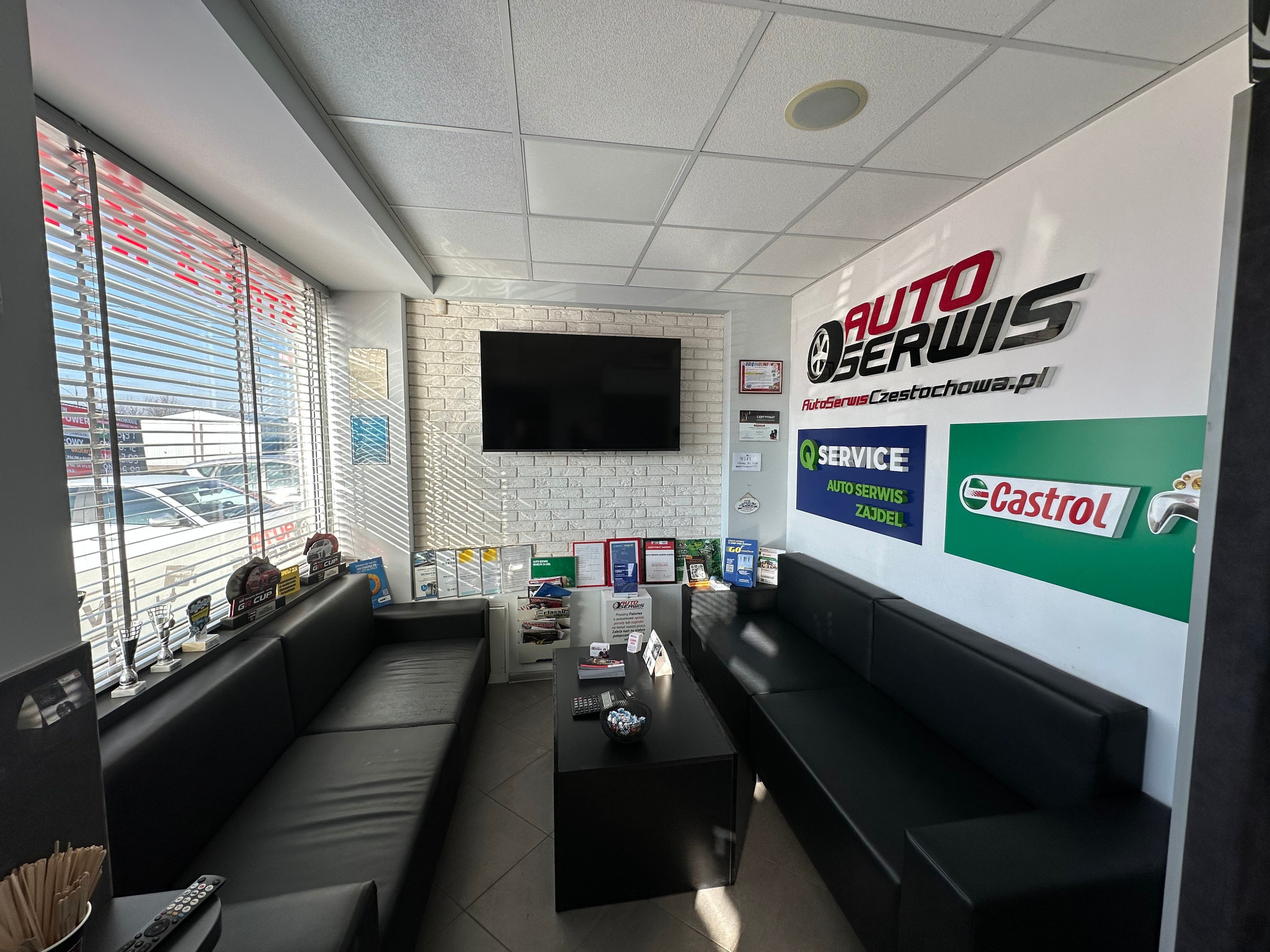 Poczekalnia auto serwisu: czarne sofy, TV na ścianie z cegły, logo serwisu, plakaty Castrol i Service. Widok na parking przez okno z żaluzjami.