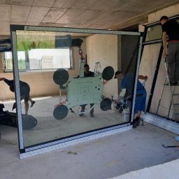 Win Bud - Montaż dużego okna. Pracownicy używają specjalistycznego sprzętu z przyssawkami do przenoszenia i instalacji. Surowy stan wykończenia wnętrza.