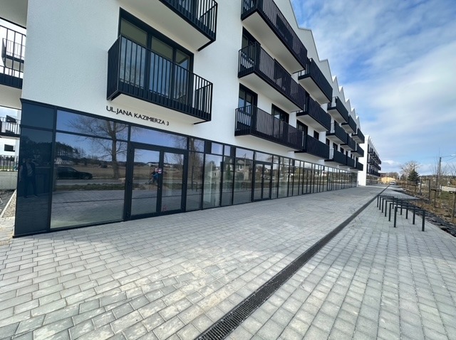 Nowoczesny budynek z czarnymi oknami i balkonami, widok z zewnątrz. Na parterze duże przeszklenia z widocznym adresem 'Ul. Jana Kazimierza 3'.