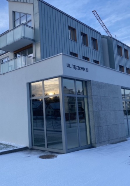 Nowoczesny budynek z dużymi, przeszklonymi oknami na parterze, odbijającymi zachodzące słońce. Widoczny adres 'Ul. Tęczowa 33' i częściowo ośnieżony teren.