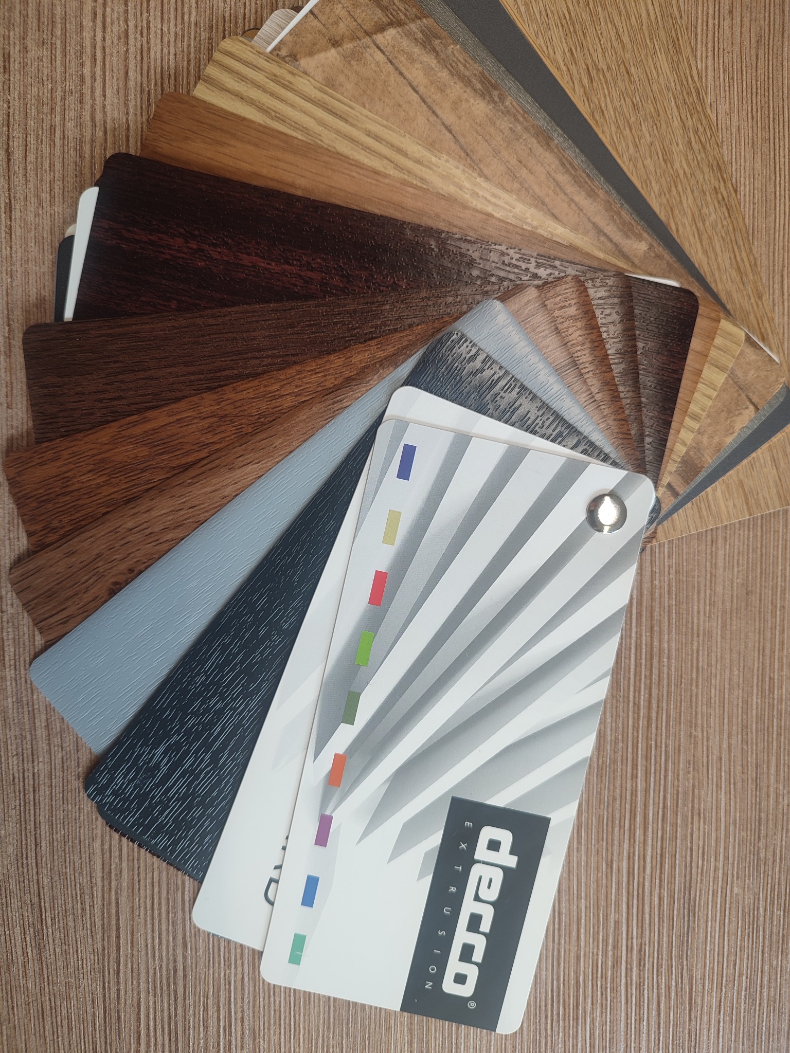 Paleta próbek kolorów i faktur paneli Decco Extrusion, ułożonych wachlarzowo na drewnopodobnym tle. Widoczne odcienie brązu, szarości i beżu, z akcentami kolorystycznymi.