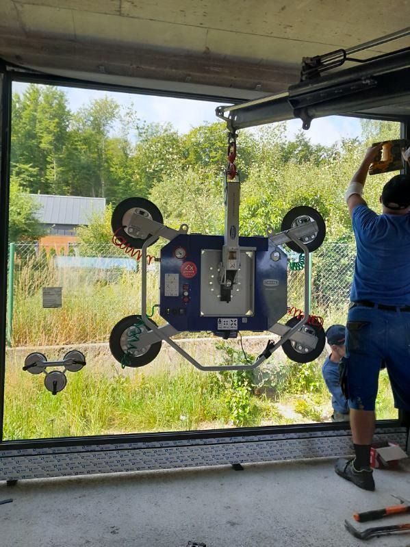 Montaż okna z użyciem przyssawki przez dwóch pracowników. Widoczne narzędzia, rama okienna i zielone tło za oknem. Okno umieszczone w surowym otworze.