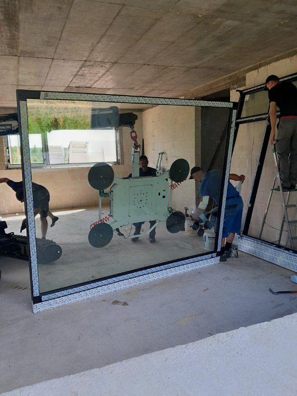 Montaż dużego okna. Pracownicy używają specjalistycznego sprzętu z przyssawkami do przenoszenia i instalacji. Surowy stan wykończenia wnętrza.