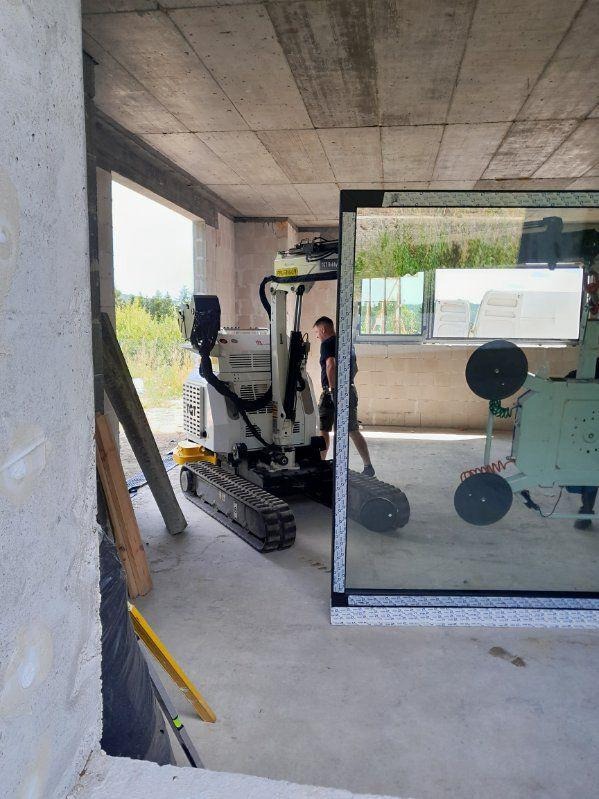 Montaż okna z użyciem robota na gąsienicach w surowym stanie budynku. Widoczne okno z szybą i pracownik nadzorujący proces.