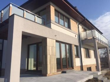 Dwukondygnacyjny dom z brązowymi oknami i szklanymi balustradami balkonów, oświetlony słońcem, na tle błękitnego nieba. Widok z poziomu gruntu.