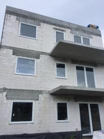 Surowy budynek w trakcie budowy z zamontowanymi białymi oknami. Widok z dołu, ukazujący konstrukcję i wstawione okna.