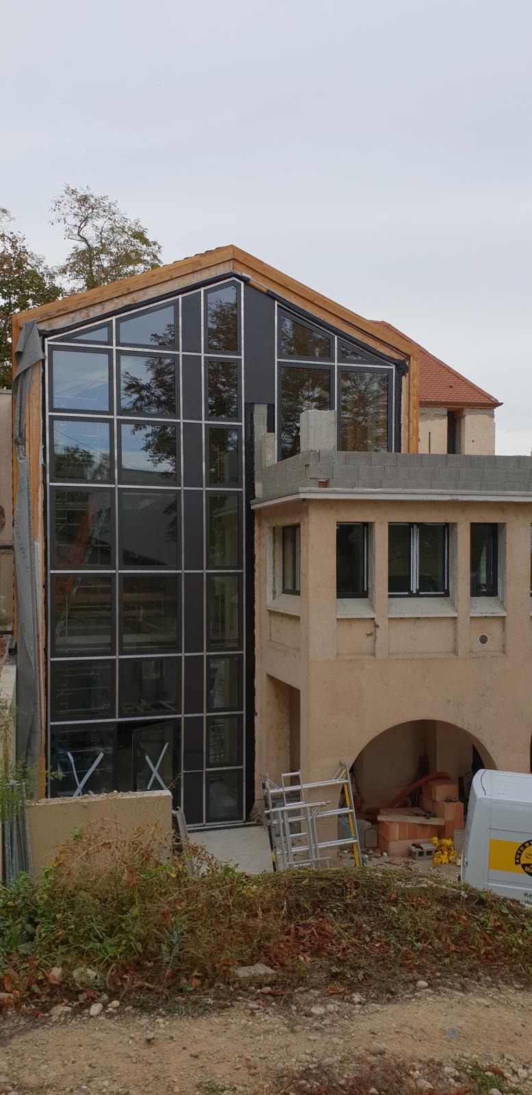 Nowoczesna fasada budynku z dużymi, kwadratowymi oknami z czarnymi ramami, odbijającymi drzewa i niebo. Widoczne rusztowania i elementy konstrukcyjne.