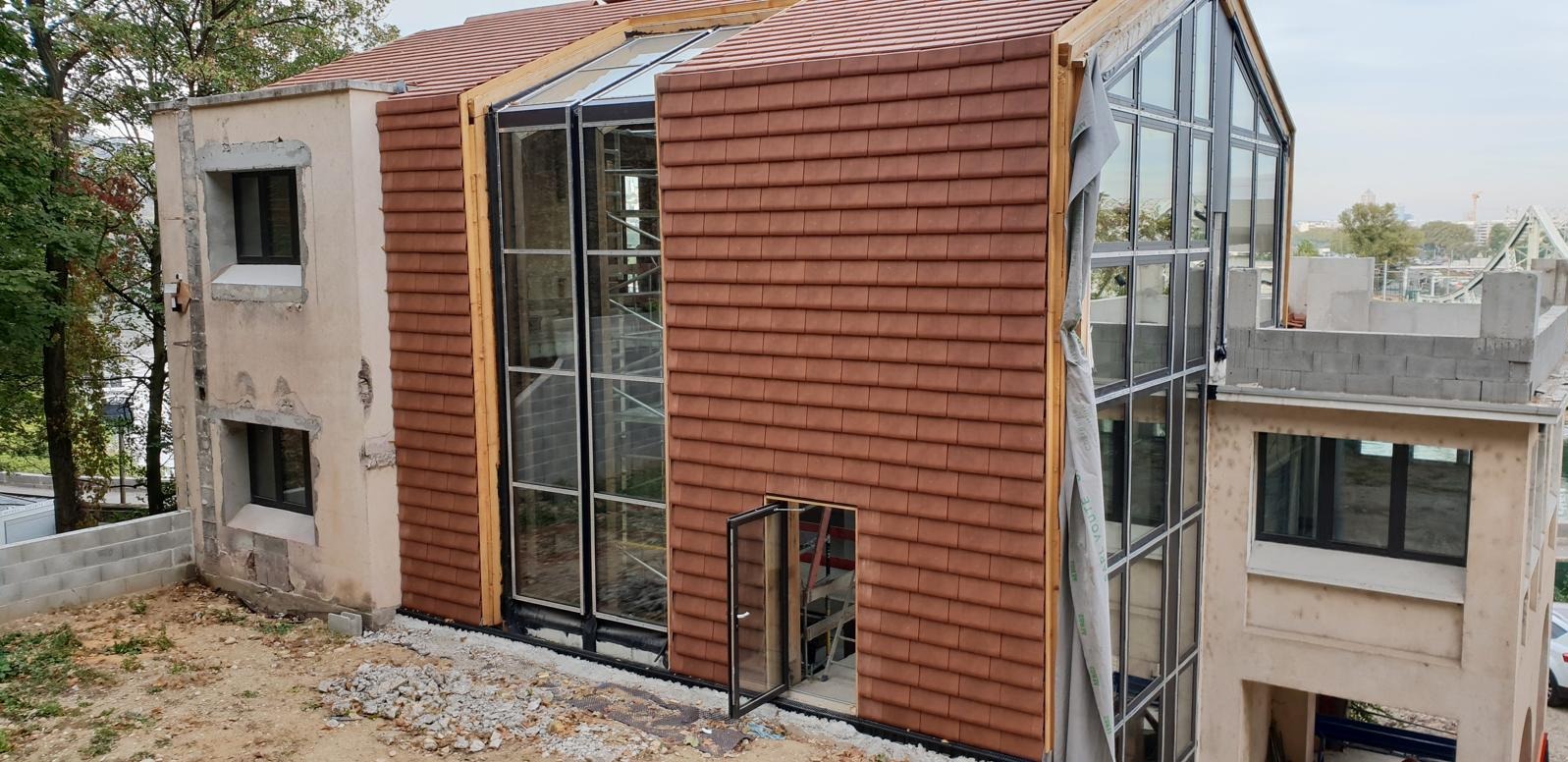 Budynek w trakcie montażu stolarki okiennej i drzwiowej, widoczne okna z czarnymi ramami i otwarte drzwi. Elewacja częściowo wykończona brązową dachówką.