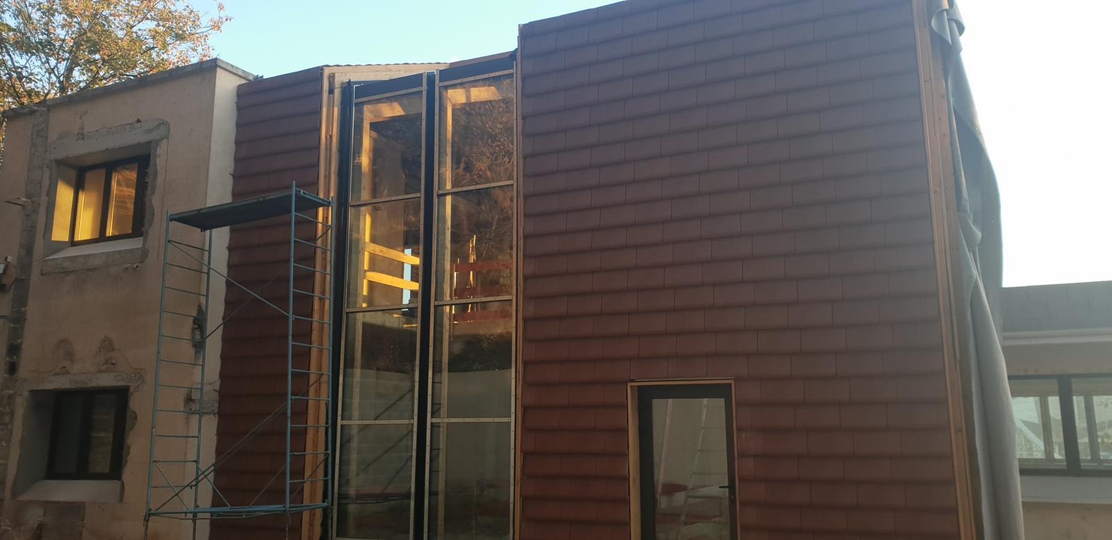 Montaż okien panoramicznych i elewacji z brązowej płytki klinkierowej na budynku w trakcie renowacji. Widoczne rusztowanie i fragmenty starej elewacji.