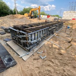 DMS BUILDING SPÓŁKA Z OGRANICZONĄ ODPOWIEDZIALNOŚCIĄ - Metalowe szalunki zbrojeniowe na piasku, przygotowane do zalania betonem. W tle koparka i elementy konstrukcji budowlanej. Widok z poziomu gruntu.