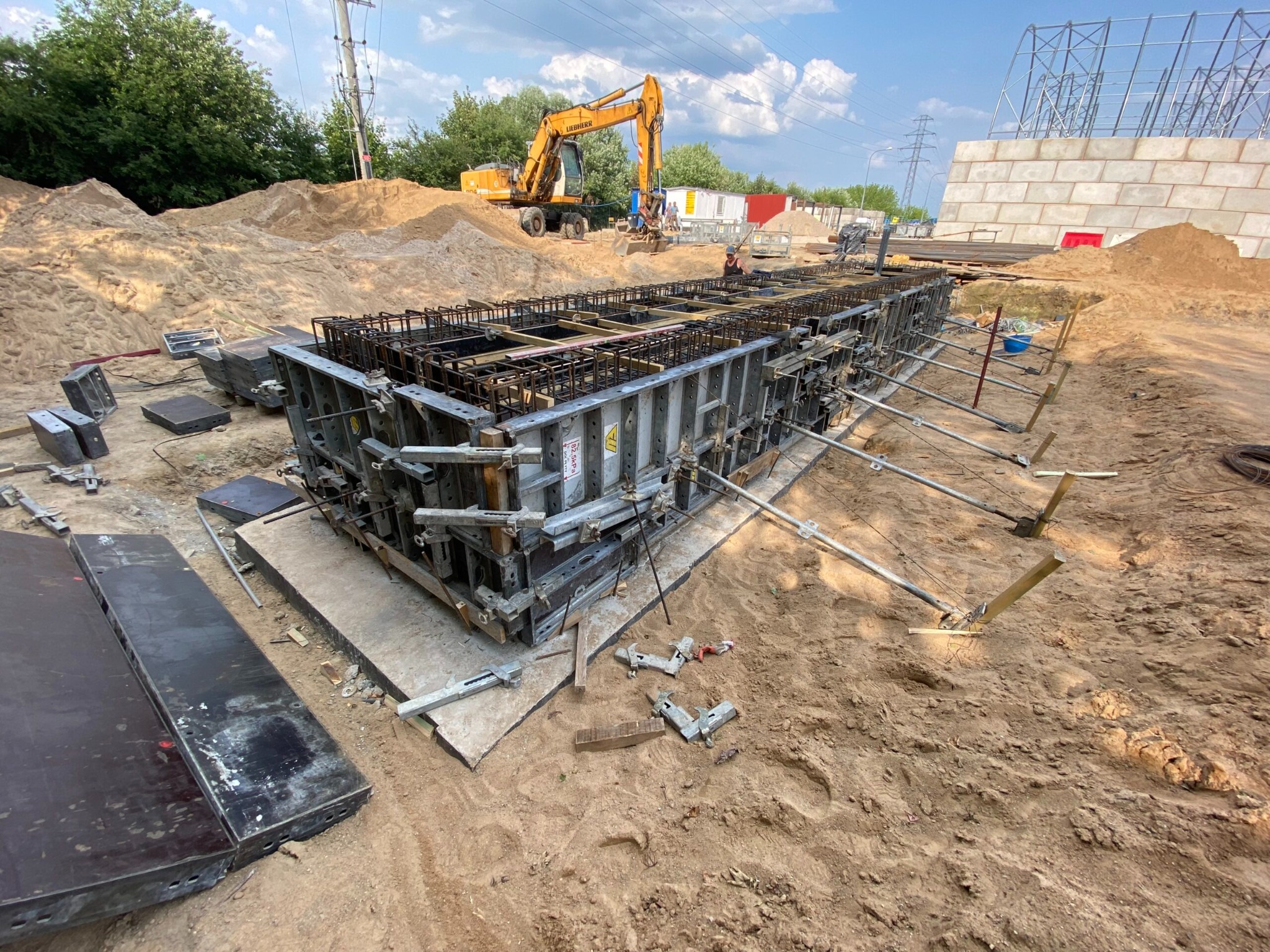 Metalowe szalunki zbrojeniowe na piasku, przygotowane do zalania betonem. W tle koparka i elementy konstrukcji budowlanej. Widok z poziomu gruntu.