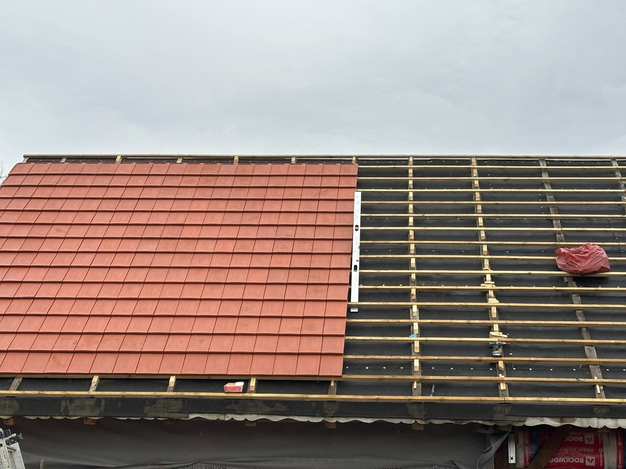 Częściowo pokryty dach z czerwoną dachówką, widoczne drewniane łaty i folia. Na niezadaszonej części leży wkrętarka, a w tle widać materiały izolacyjne.