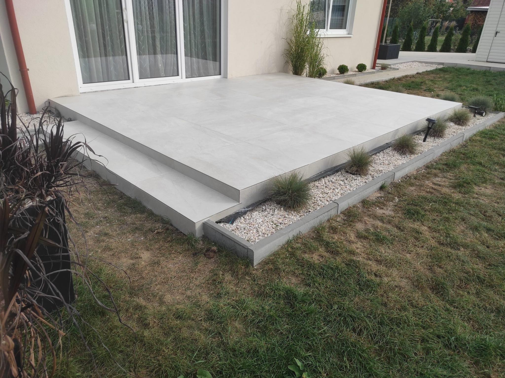 Wykończony taras z jasnych płyt, z dwoma stopniami i obrzeżem z kamyków oraz niskich roślin. Oświetlenie solarne wzdłuż krawędzi. Widok na dom i ogród.