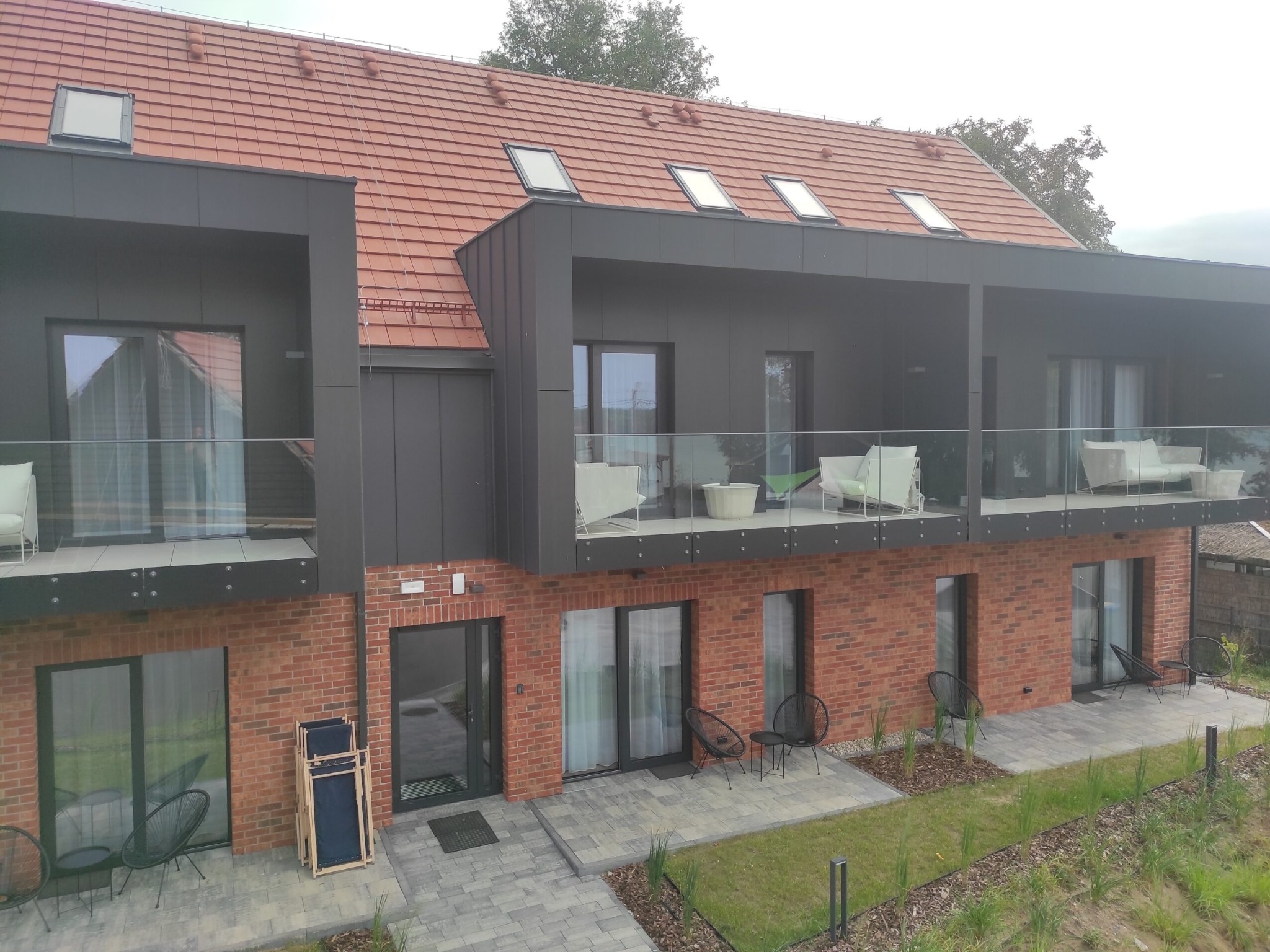 Nowoczesny budynek z balkonami i czerwoną dachówką. Elewacja z cegły i szarych paneli. Widoczne okna dachowe i meble balkonowe.