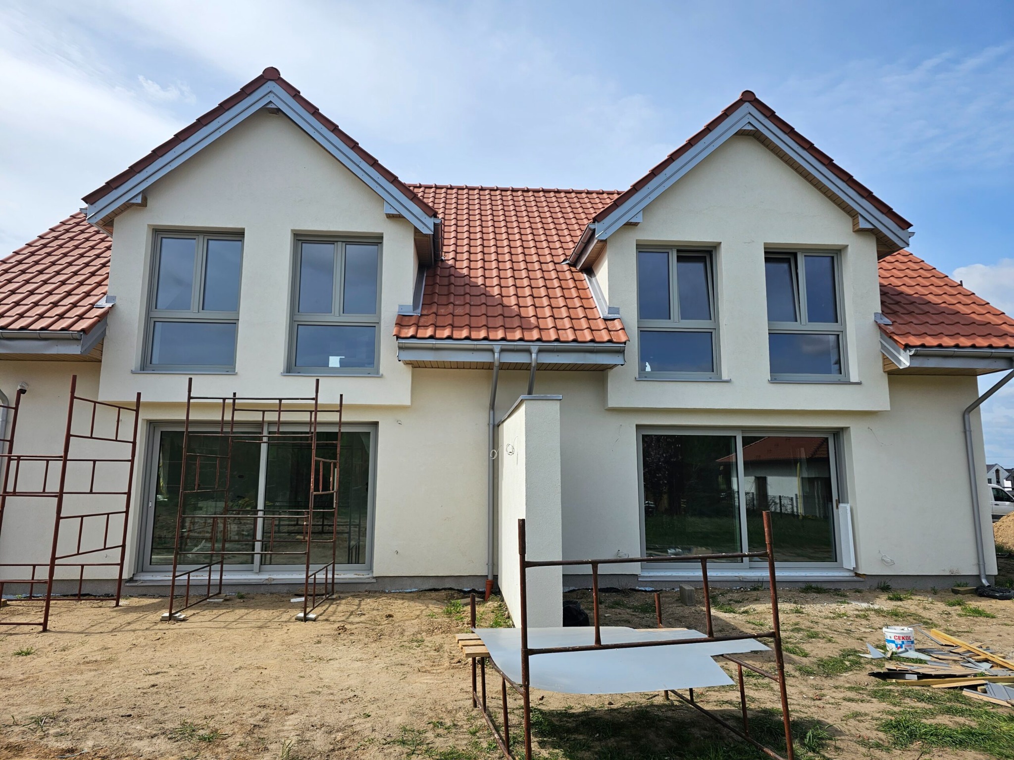 Dwukondygnacyjny dom z jasną elewacją i czerwoną dachówką w trakcie budowy, widoczne rusztowania i narzędzia. Ujęcie w słoneczny dzień z błękitnym niebem.